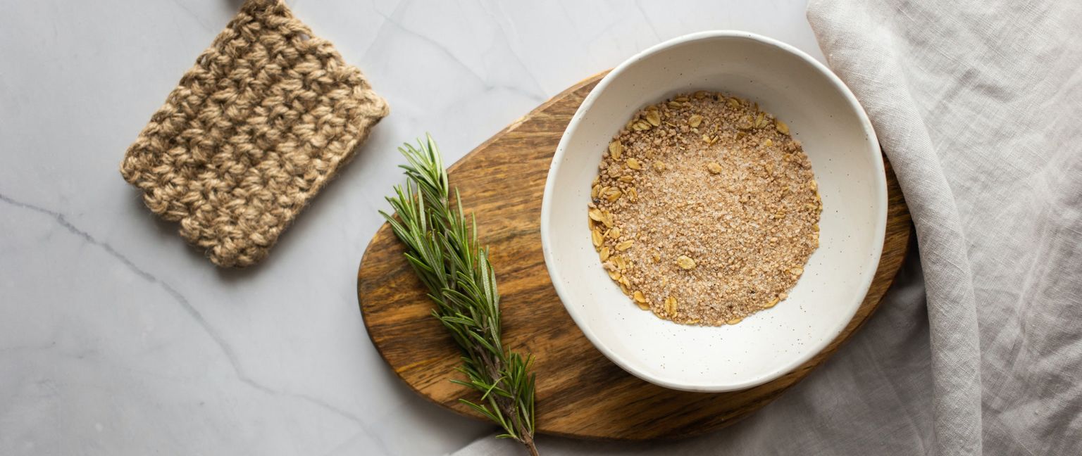 A top-down shot of a bowl containing a mixture of grain and sugar on a wooden cutting board, next to a sprig of rosemary and hemp dishcloth. A gray linen napkin is draped to the side on a white marble surface.