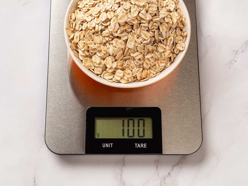 A white bowl filled with old fashioned oats sits on a digital food scale. The scale reads 100 grams. The background is white with grey marbling.