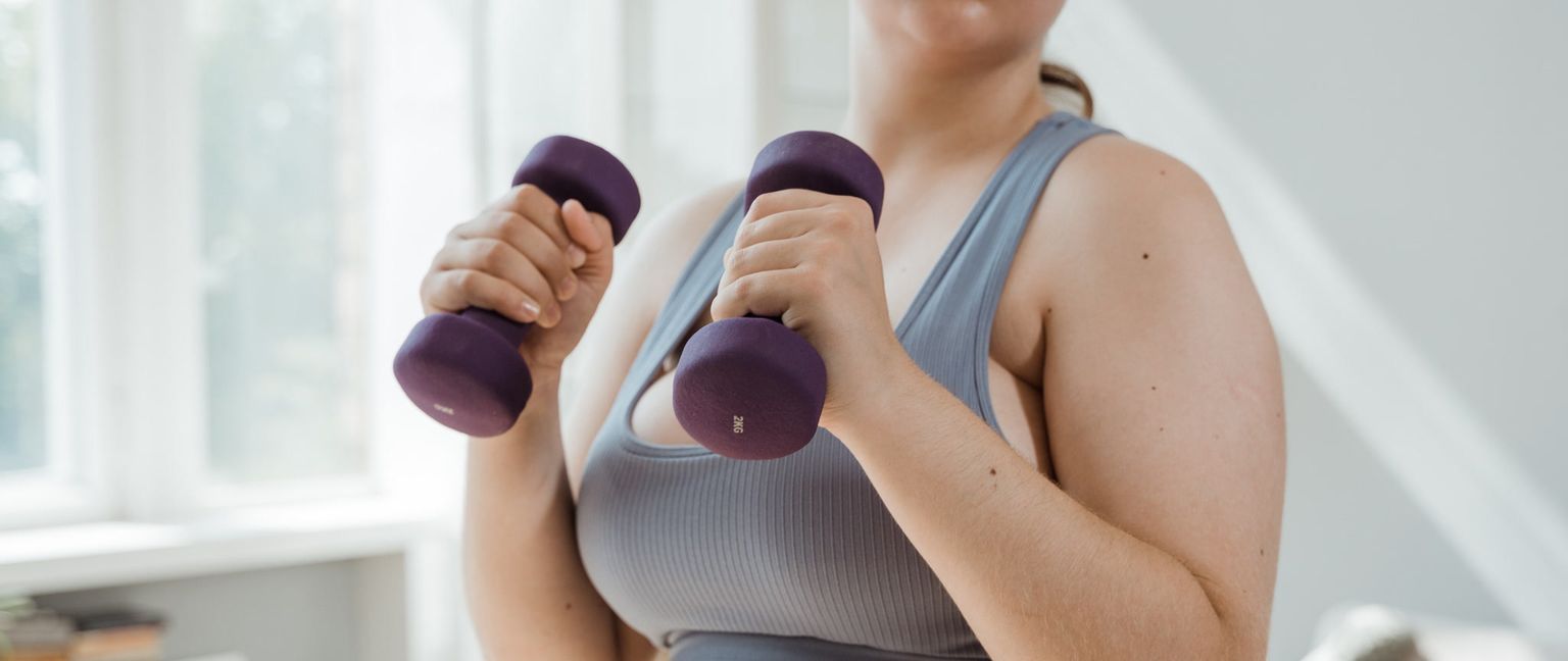 A woman is shown from the chest up, holding 2kg dumbbells in each hand during a workout.