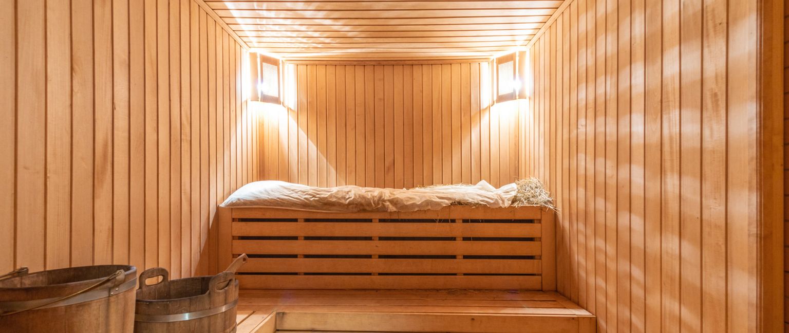 Interior view of a wooden sauna illuminated by two wall-mounted electric lights, showing benches and buckets.