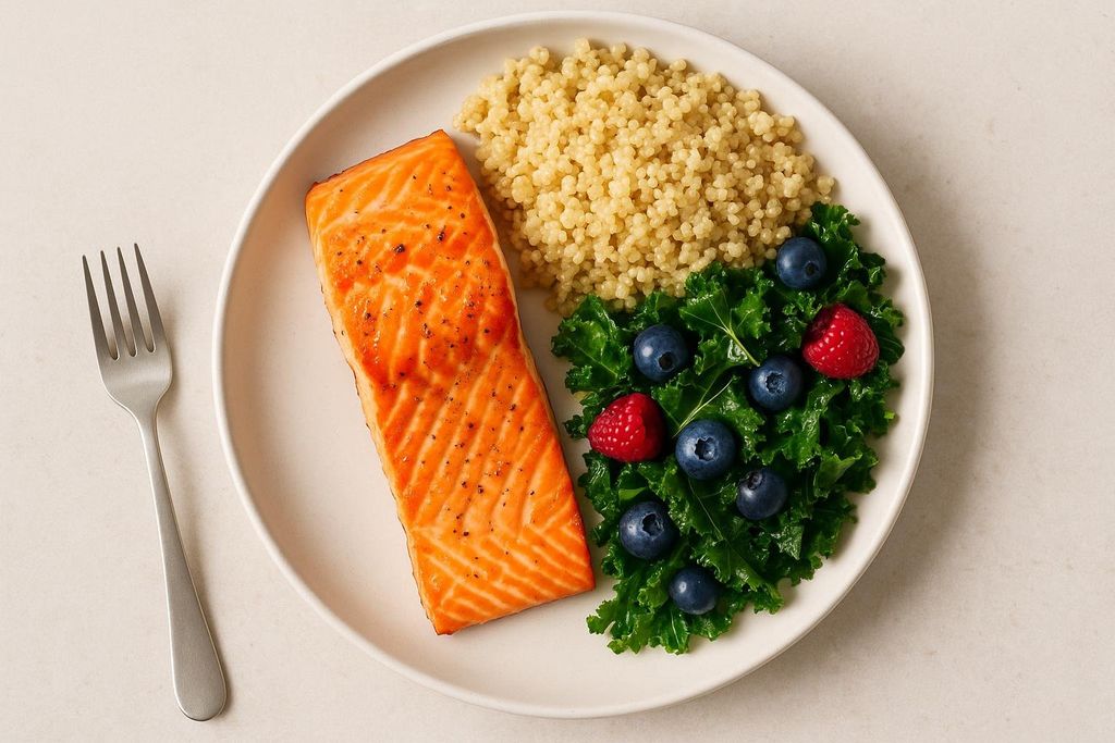 A white plate contains a meal with a large piece of cooked salmon, a serving of quinoa, and a side of kale topped with blueberries and raspberries. A silver fork is next to the plate.
