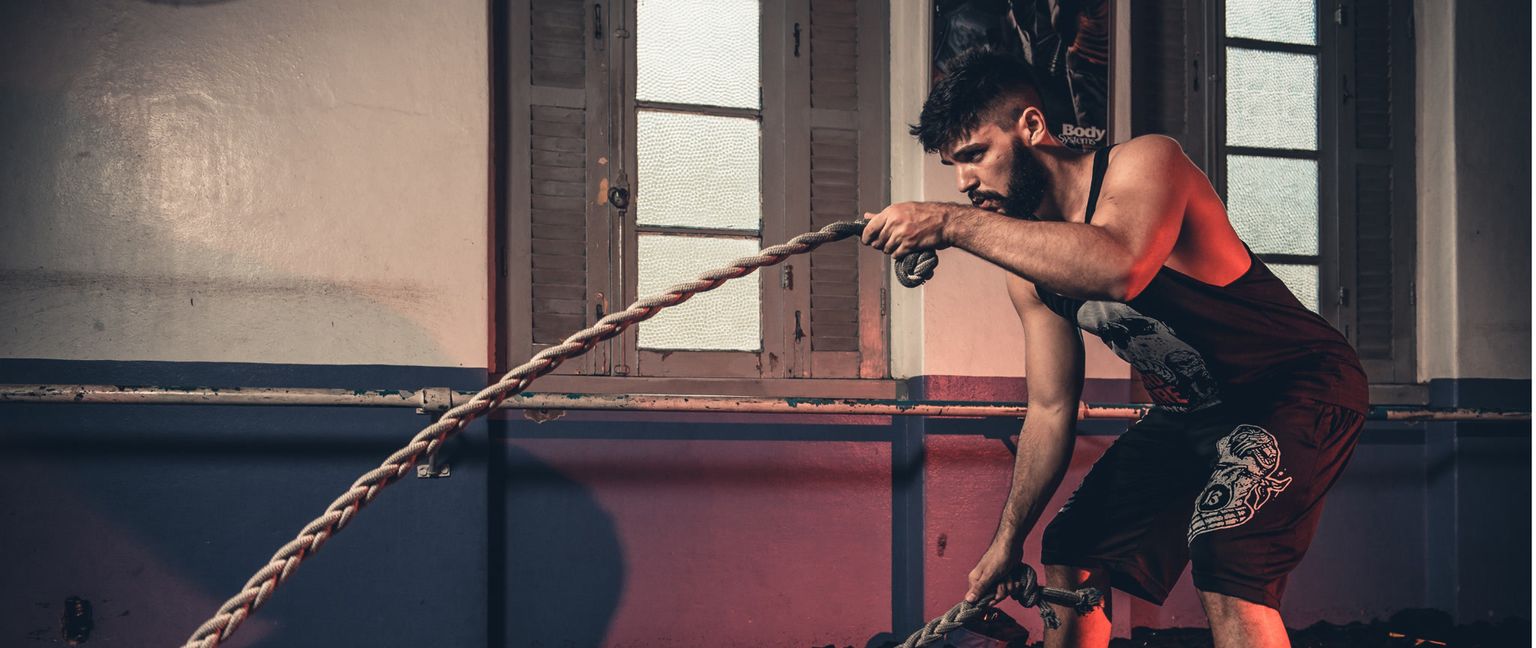 A man in a black tank top and shorts uses battle ropes in a gym.