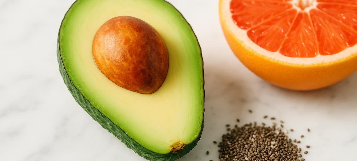 A close-up shot of a halved avocado with its pit on a marble surface. Next to it, a half-cut grapefruit and a small pile of chia seeds.