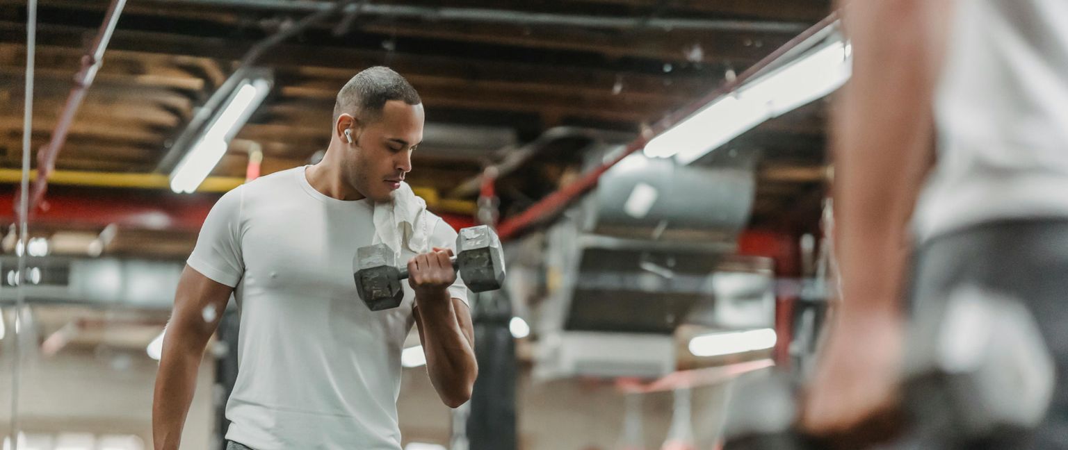 A man in a white t-shirt and earbuds curls a dumbbell in a gym, with his reflection visible in a mirror.