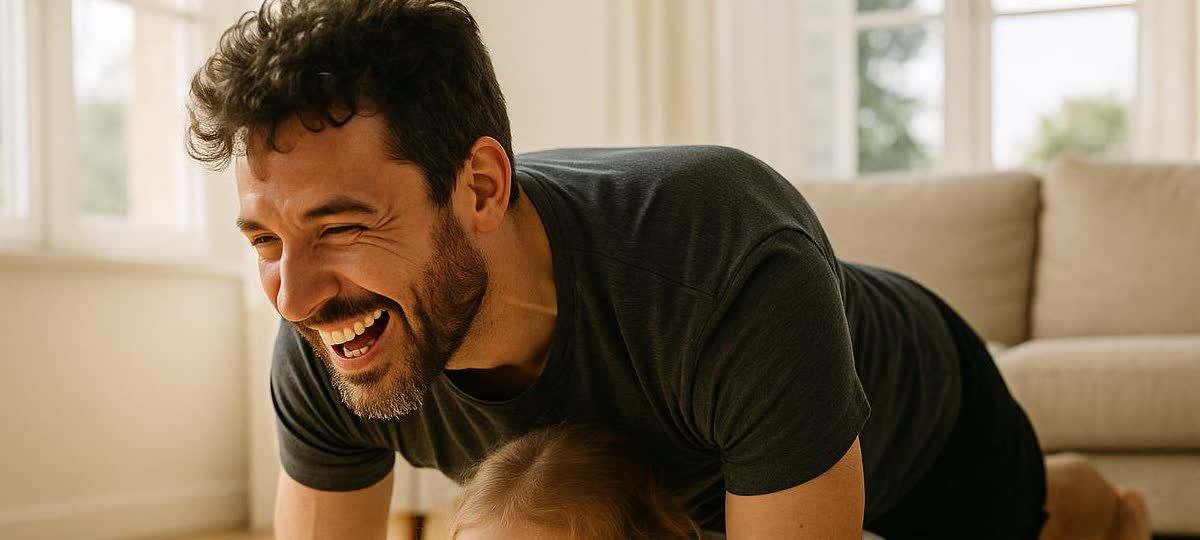 A father and his young daughter exercising playfully at home in a bright living room. The father is laughing and smiling at the camera as he appears to be in a plank position over his daughter.