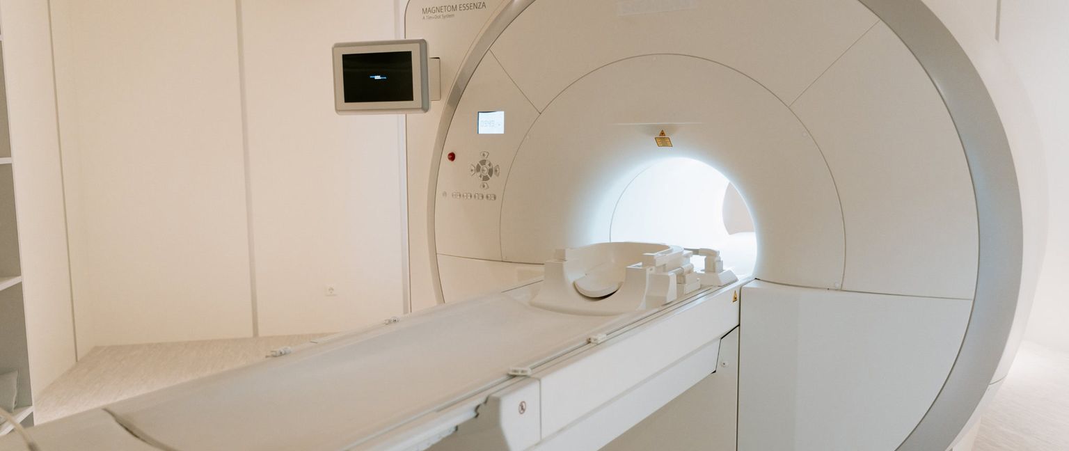 A white large Magnetic Resonance Imaging (MRI) machine inside a brightly lit, sterile medical room. A bed for the patient slides into the large circular opening of the machine.
