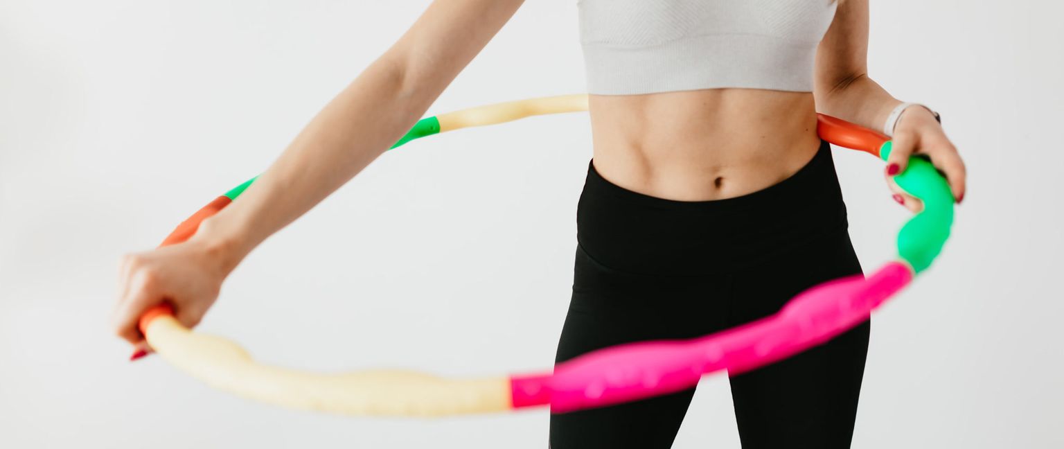 A woman's torso and arms as she uses a colorful hula hoop for exercise. The focus is on her toned abdominal muscles and the movement of the hoop.