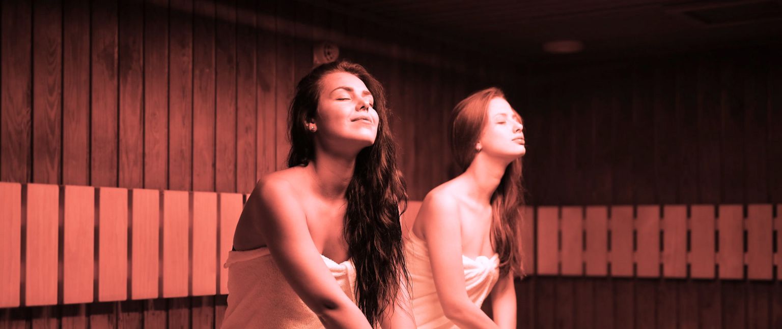 Two young women wrapped in towels are relaxing in a sauna, with their eyes closed and heads tilted back.