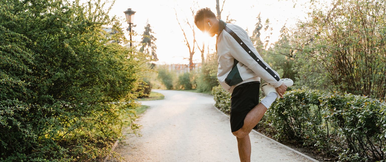 A man stretches his thigh by pulling his foot towards his backside in a park at sunset.