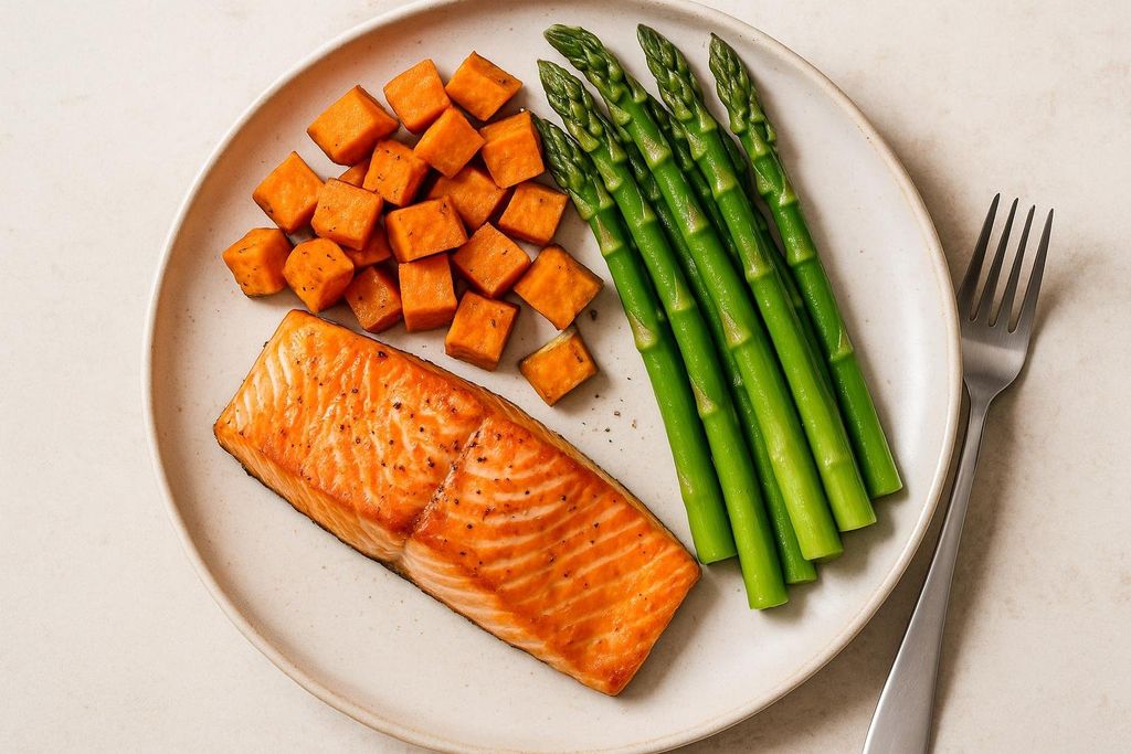 A balanced, high-protein meal on a plate featuring a cooked salmon fillet, cubed sweet potatoes seasoned with pepper, and vibrant green asparagus spears. A fork is visible on the right side of the plate.