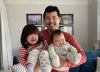 A father smiles broadly while holding his crying toddler daughter on his left and his smiling infant son on his right. The father is wearing a red shirt, the toddler has short dark hair and is wearing a red and white striped shirt and cloud-patterned leggings, and the baby is wearing a grey and white cloud-patterned onesie.