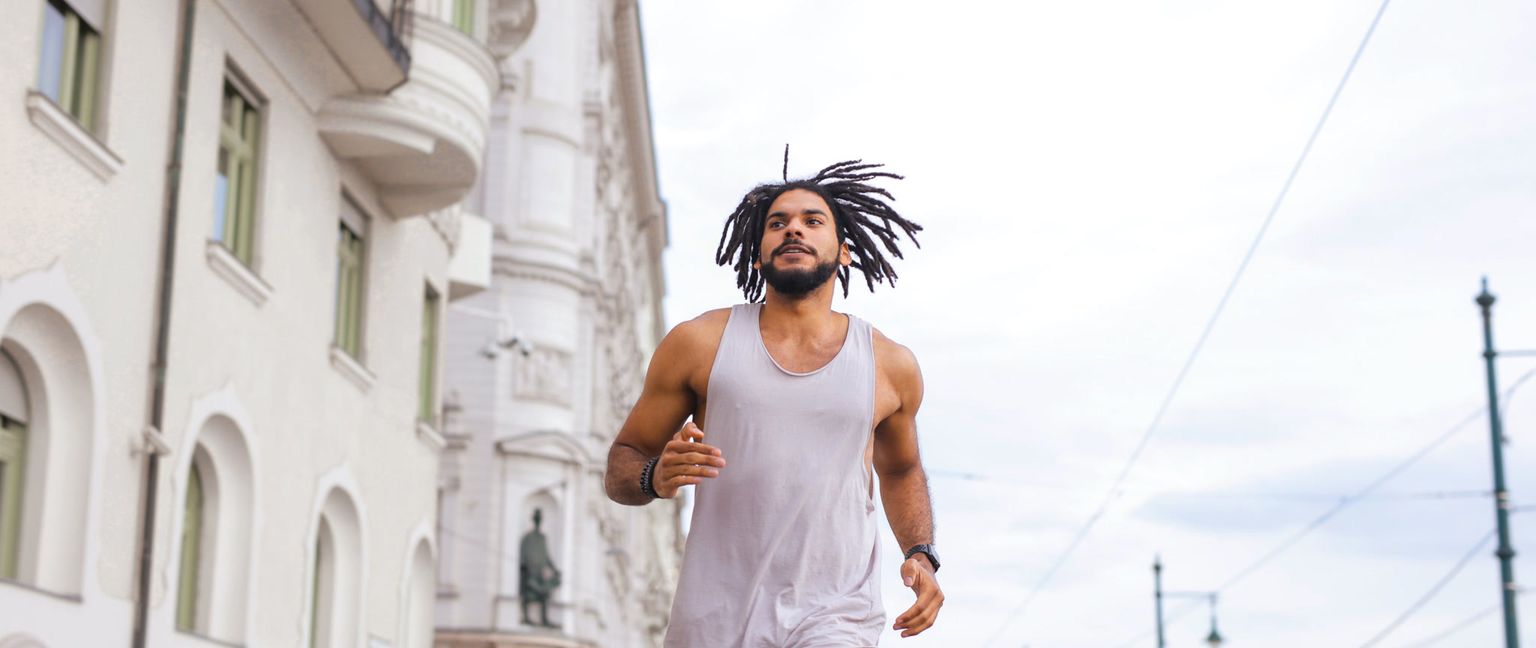 A man with dreadlocks and a tank top running with buildings in the background.