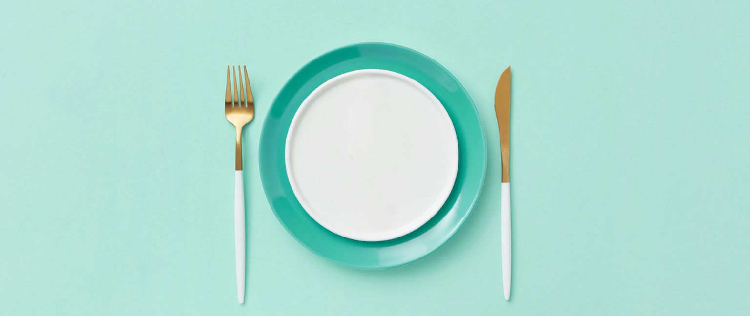 An empty white plate sits on a larger teal plate with a gold knife and fork on a light green background.