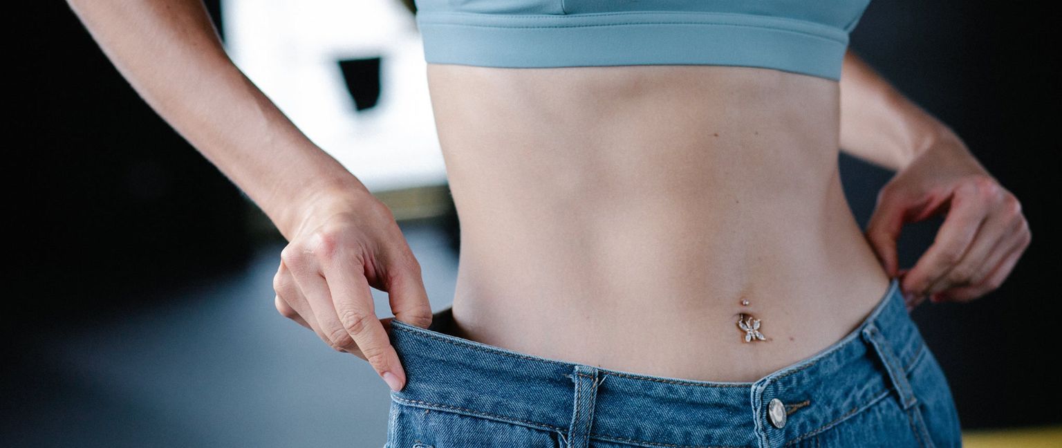 A close-up of a woman's midsection, holding her loose-fitting jeans away from her waist to show significant weight loss.