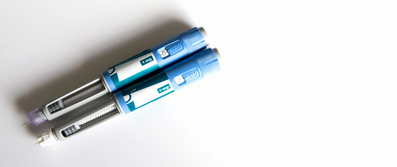 Two blue and white GLP-1 injector pens with needles visible, laying on a white background.
