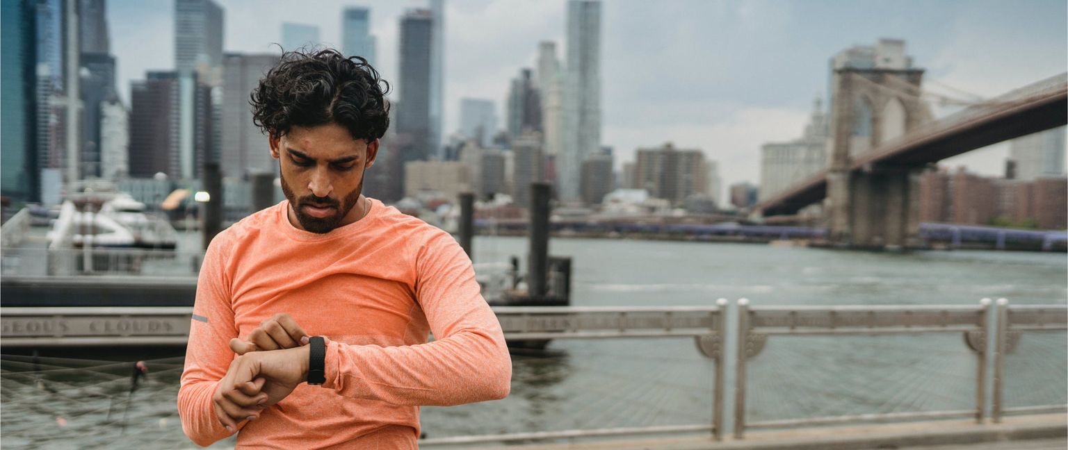 A man checks his smartwatch with a city skyline and Brooklyn Bridge in the background.