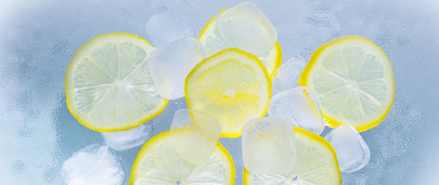 Several bright yellow lemon slices and clear ice cubes are scattered across a light blue, textured background. The light reflects off the ice and the surfaces of the lemon slices.