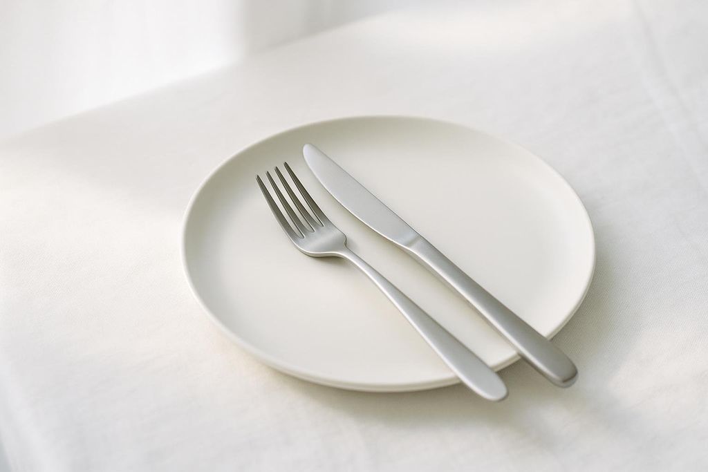 An empty white dinner plate with a silver fork and knife resting on it. The plate is positioned on a white tablecloth, and soft light casts subtle shadows.
