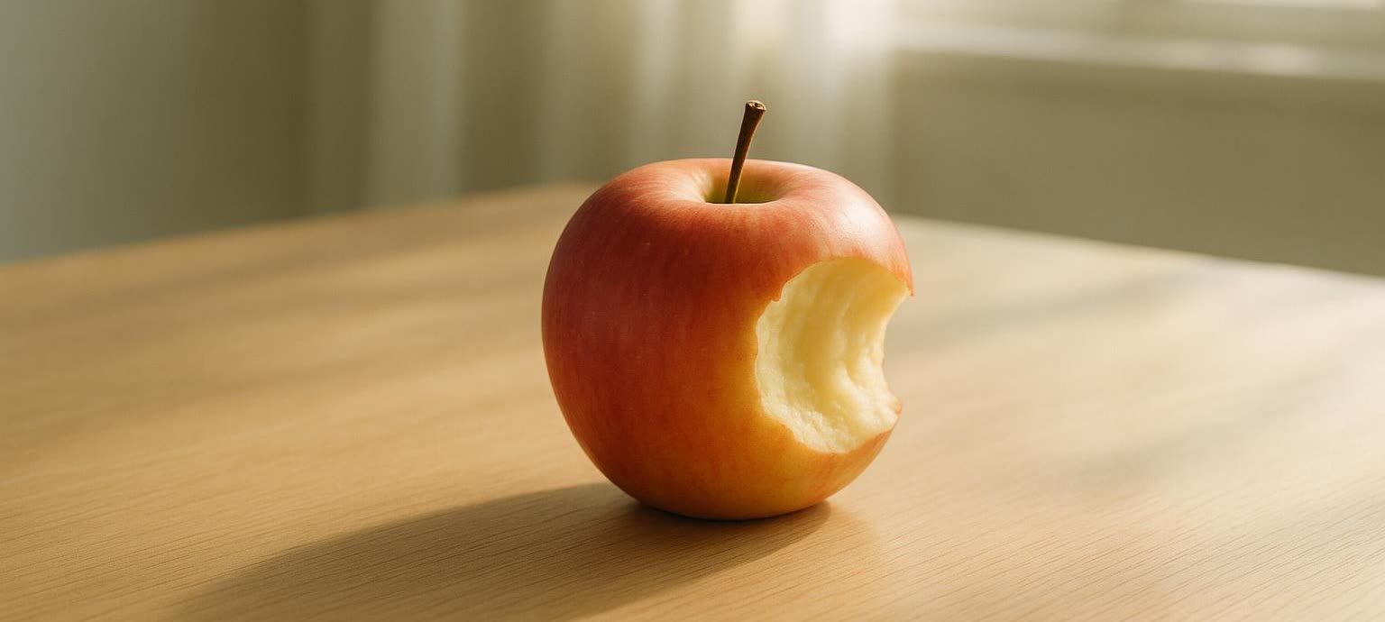A single reddish-yellow apple sits on a light brown wooden surface. A significant bite has been taken out of the apple, exposing its white, crisp interior.
