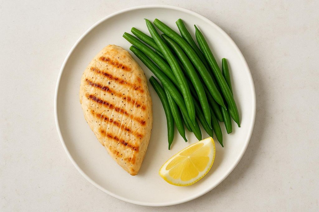 A white plate holds a grilled chicken breast, a pile of bright green beans, and a small wedge of lemon.