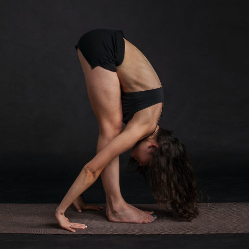 A woman in black yoga clothes performing a standing forward fold with her legs together, arms wrapped around her calves with hands touching the mat behind her heels, and forehead touching her knees. Her long, dark hair hangs towards the floor. She is on a yoga mat against a dark background.