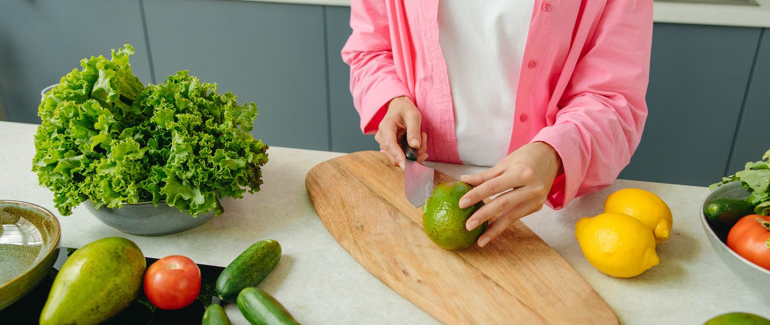 A woman in a pink shirt is cutting an avocado on a wooden cutting board. Other vegetables, including lettuce, tomatoes, cucumbers, and lemons are on the counter around the cutting board.