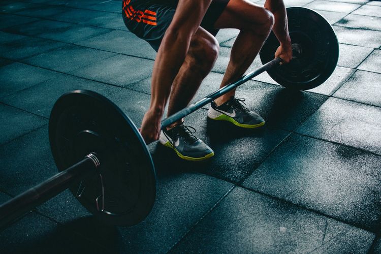 A low angle view of a person in shorts and athletic shoes bending down with a barbell, preparing to lift it.