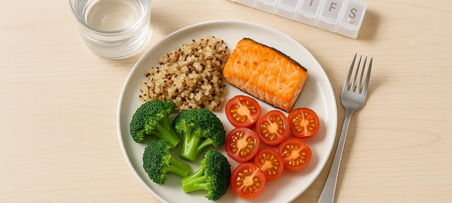A plate featuring a balanced meal with pan-seared salmon, quinoa, steamed broccoli florets, and halved cherry tomatoes. A glass of water and a pill organizer are also visible on the light wood table.