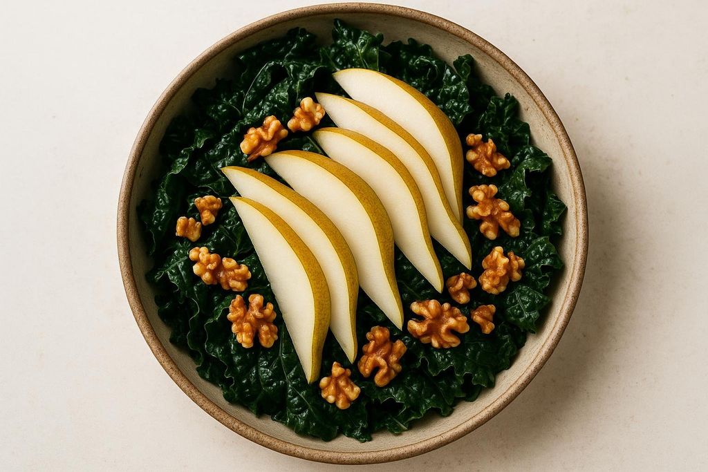 An overhead view of a vibrant kale salad in a bowl, garnished with five elegant slices of ripe pear and scattered whole walnuts. The deep green kale contrasts beautifully with the light yellow-green pears and golden-brown walnuts.