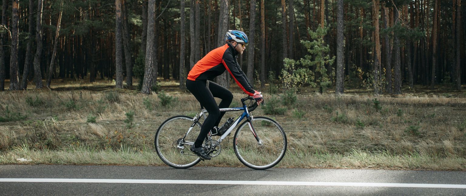 A person wearing a helmet and orange jacket cycles on a road that runs alongside a forest.