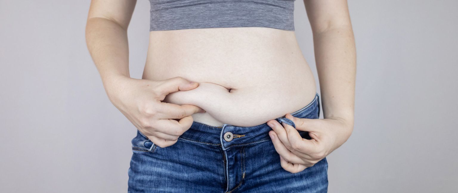 A woman with her hands on her stomach is pinching a roll of belly fat. She is wearing a grey sports bra and blue jeans.