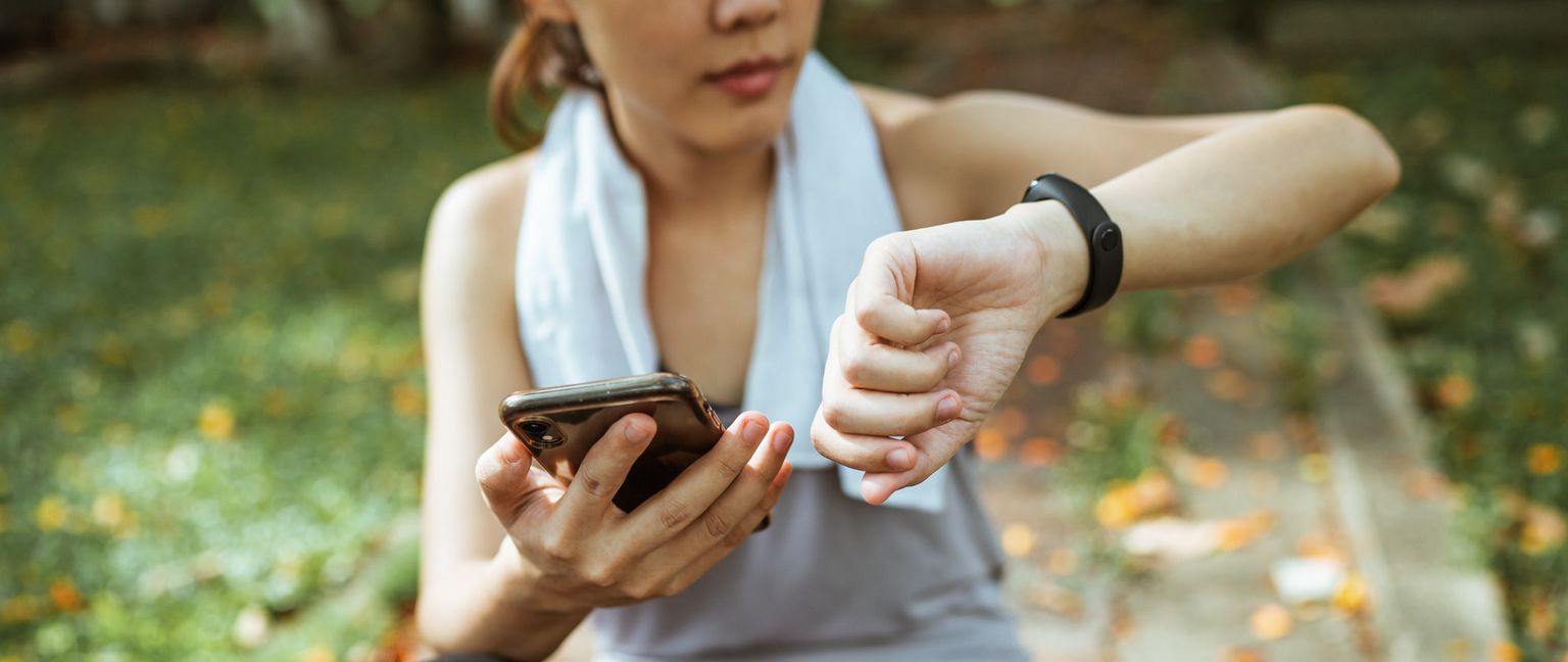 A woman wearing a fitness tracker looks at her watch while holding her phone. A towel is around her neck. She appears to be outdoors in a park or natural setting.