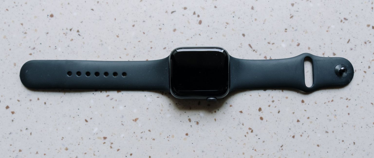 An Apple Watch with a dark gray band and black screen is displayed horizontally on a speckled white surface.