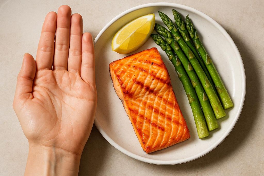 A human hand held up beside a plate showing a grilled salmon fillet, a lemon wedge, and steamed asparagus. The salmon portion is approximately the size of the palm, demonstrating a visual guide for protein intake.