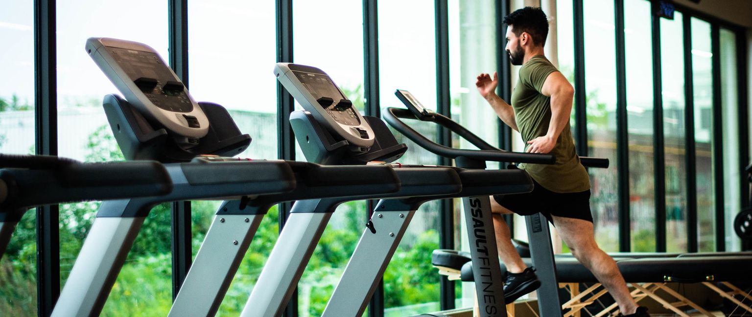 A man wearing a green t-shirt and black shorts runs on a treadmill in a gym with large windows.