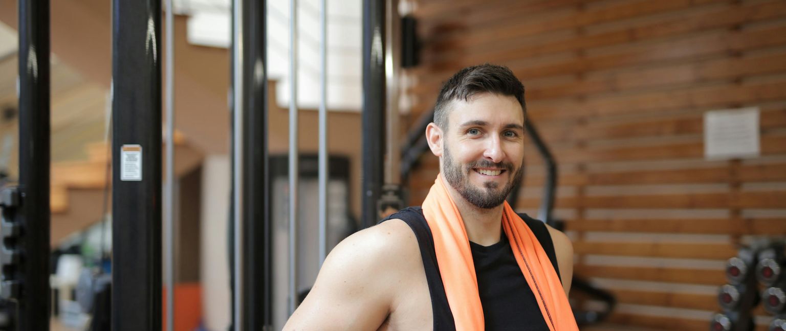 A smiling man in a black workout shirt with an orange towel around his neck stands in a gym.