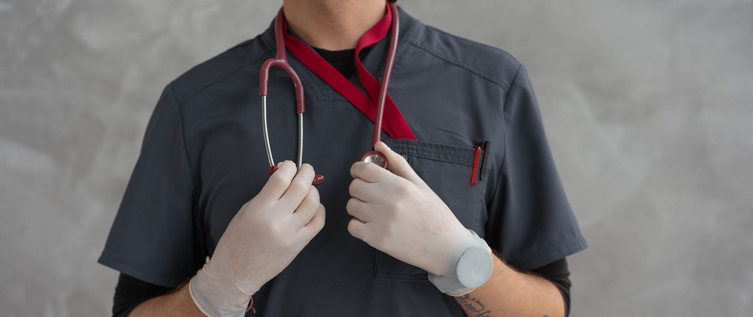 A doctor in dark grey scrubs and white gloves holds a red stethoscope in their hands.
