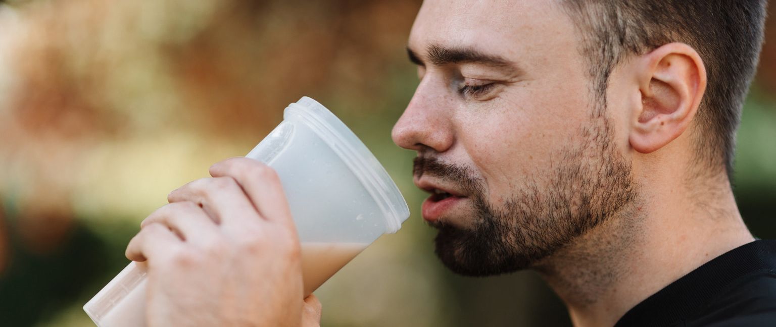 Close-up of a man drinking a protein shake from a clear plastic bottle. He is wearing a black shirt and has a beard.