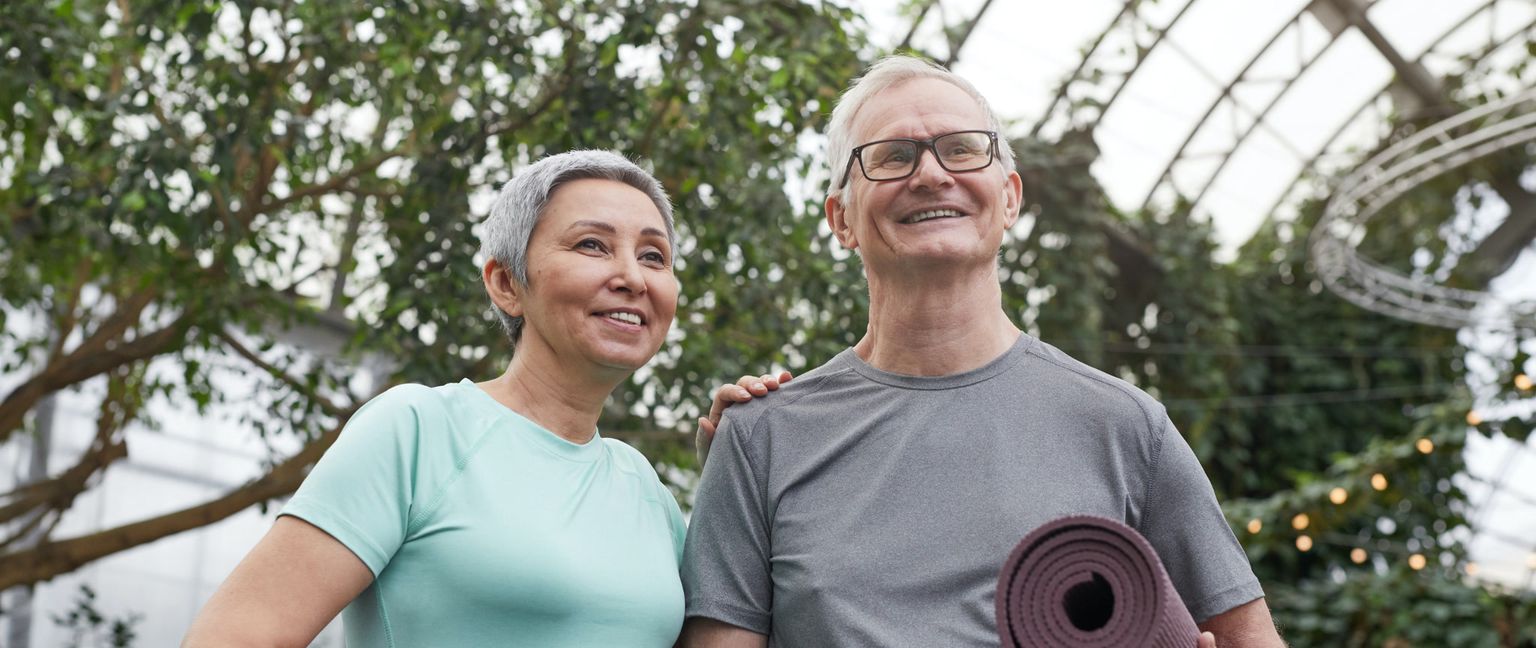 An older man and woman smile and look up, with trees and a metal structure behind them. The man holds a yoga mat.