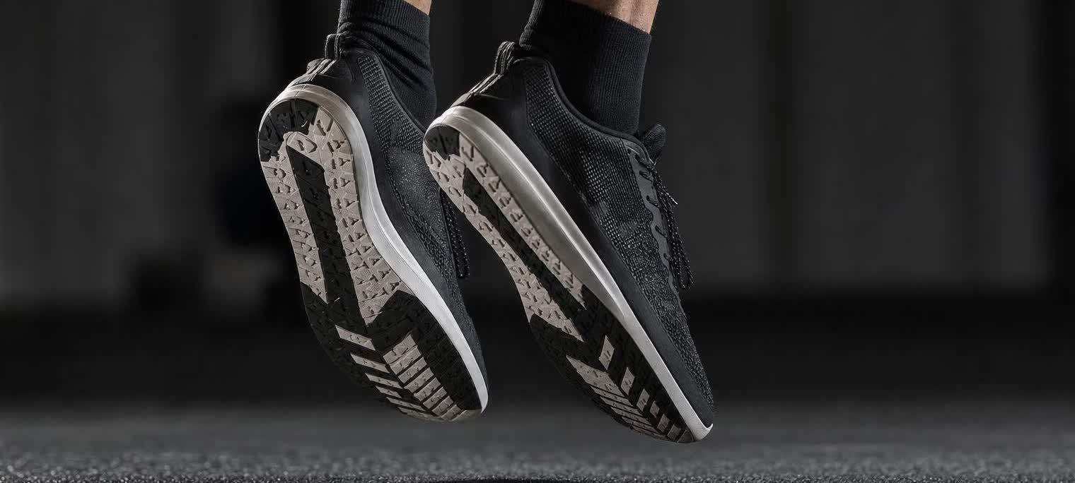 Close-up of a pair of black athletic shoes, with white soles and intricate tread patterns, suspended in mid-air just above a dark, textured gym floor. The shoes are oriented as if during a jump, with the heels slightly elevated.
