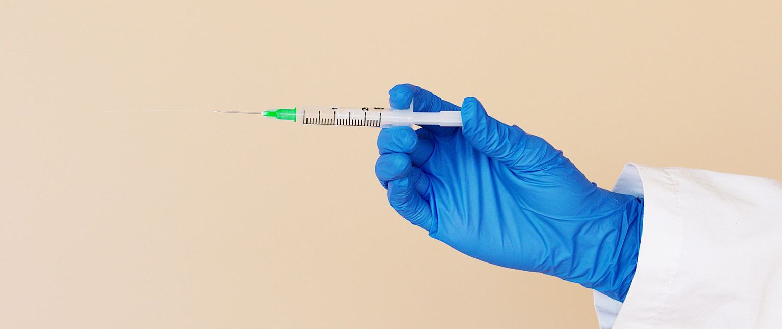 A person wearing a white coat and blue glove holds a syringe with a needle on a neutral background.