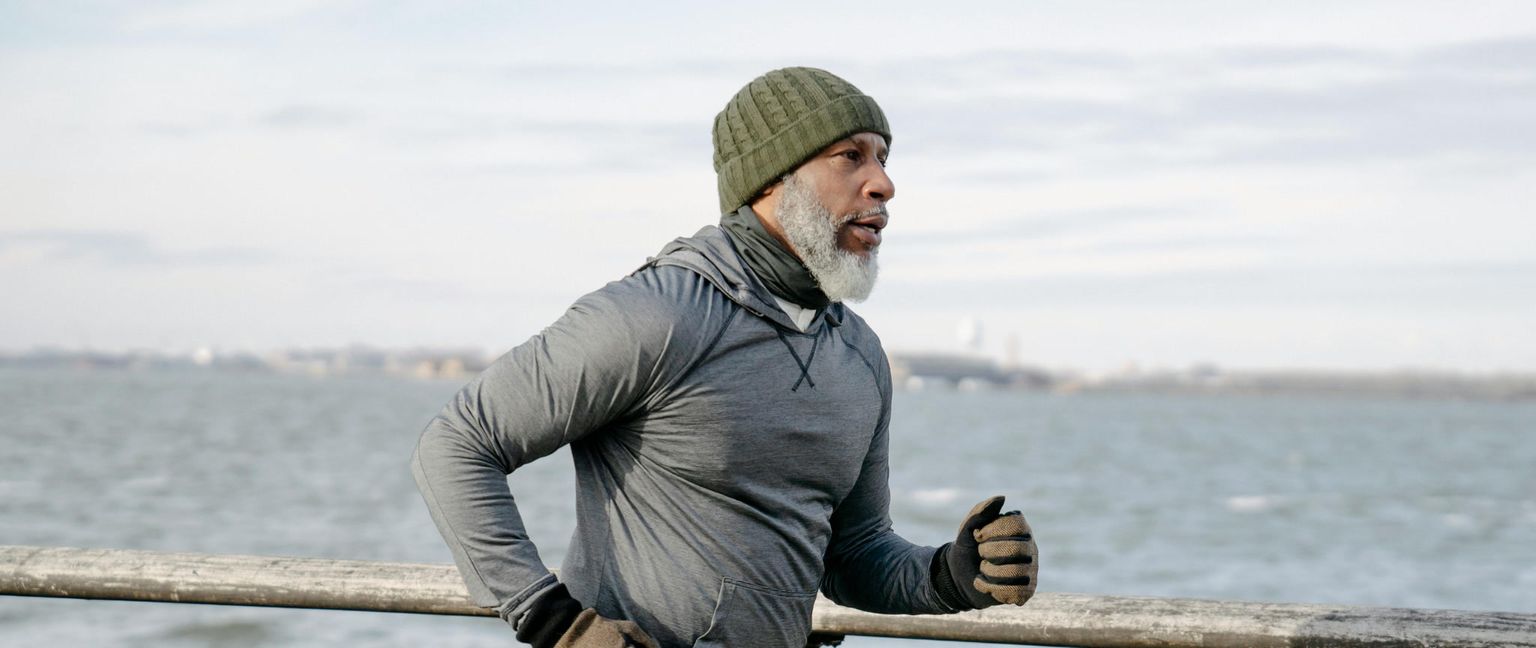 A man with a salt-and-pepper beard, wearing a beanie, grey hoodie, and gloves, runs outdoors along a railing near the water.
