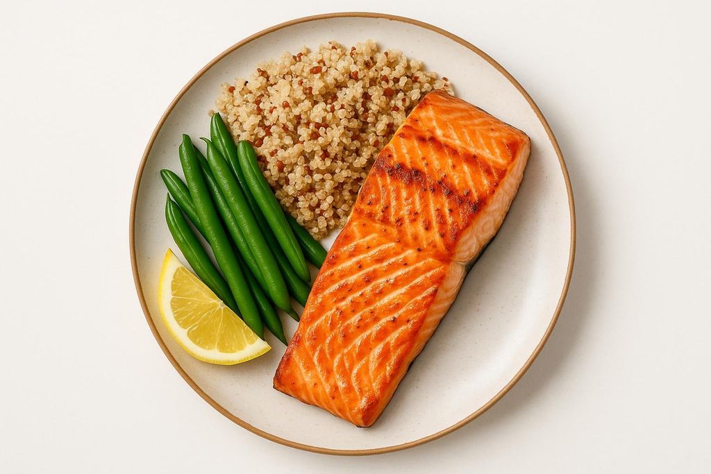 A healthy meal on a light-colored plate, featuring a perfectly grilled salmon fillet, a serving of fluffy quinoa, a side of vibrant green beans, and a wedge of fresh lemon. The salmon has prominent grill marks and a golden-orange hue, indicating it's well-cooked and flavorful.