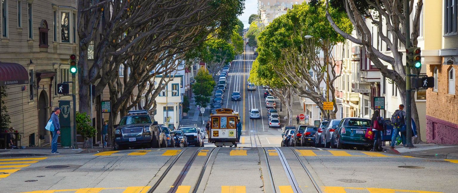 A scenic view of a San Francisco street with a cable car, steep hill, and trees lining the road.