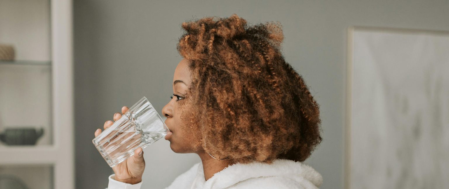 A woman with curly brown hair viewed from the side, drinking a glass of water.