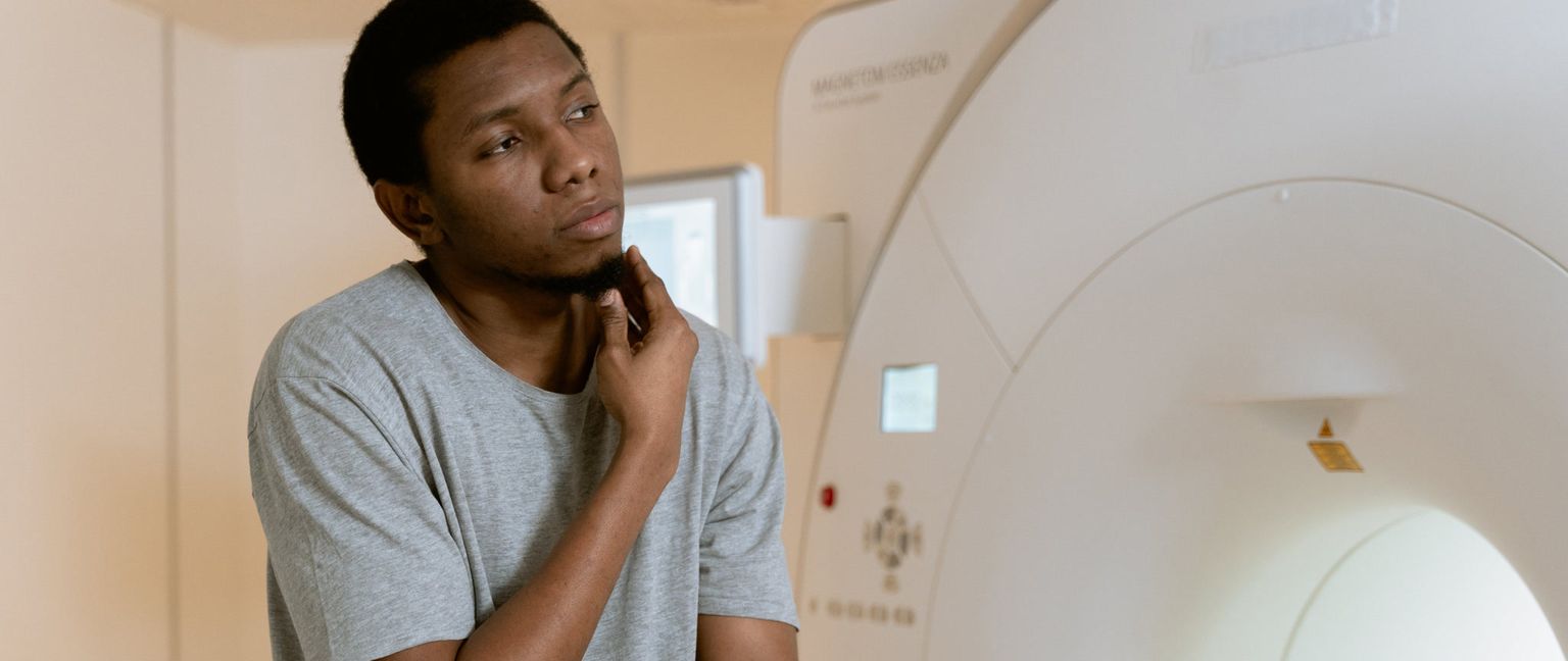 A young man in a grey t-shirt sits near a large white MRI machine, looking thoughtful and touching his chin. The scanner has a bright light emanating from its opening.