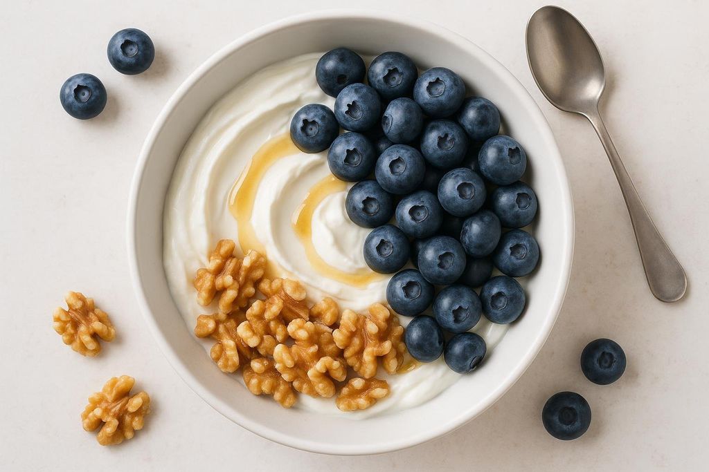 A white bowl filled with creamy yogurt, topped with fresh blueberries, walnuts, and a drizzle of honey. A silver spoon rests beside the bowl on a light background.