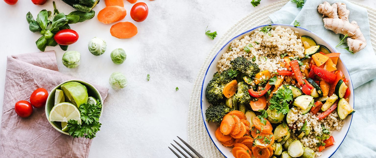 A vibrant bowl filled with quinoa, broccoli, sliced carrots, zucchini, and bell peppers, surrounded by cherry tomatoes, brussels sprouts, and lime wedges.