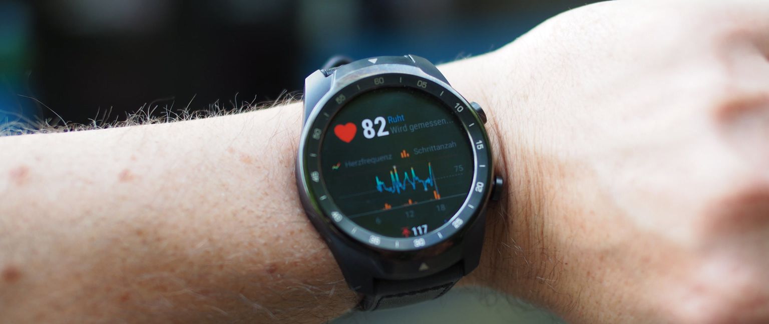 A close-up of a black fitness watch on a person's wrist, displaying a heart rate of 82bpm and a line graph showing heart rate over time. The watch also shows 'Schrittanzahl' and a value of 117, likely representing steps counted.
