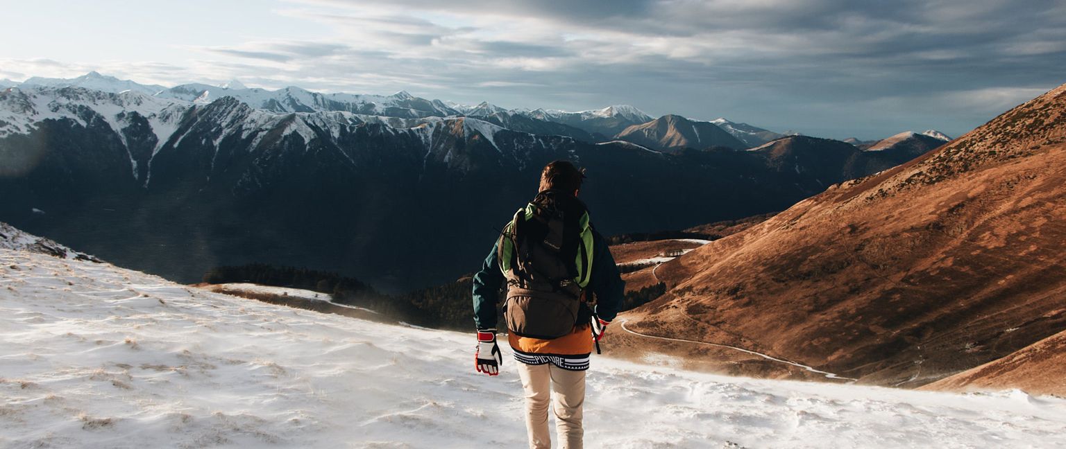 A man with a backpack hikes on a snowy mountain ridge, overlooking lower brown mountains and a distant river, with snow-capped peaks in the background under a cloudy sky.